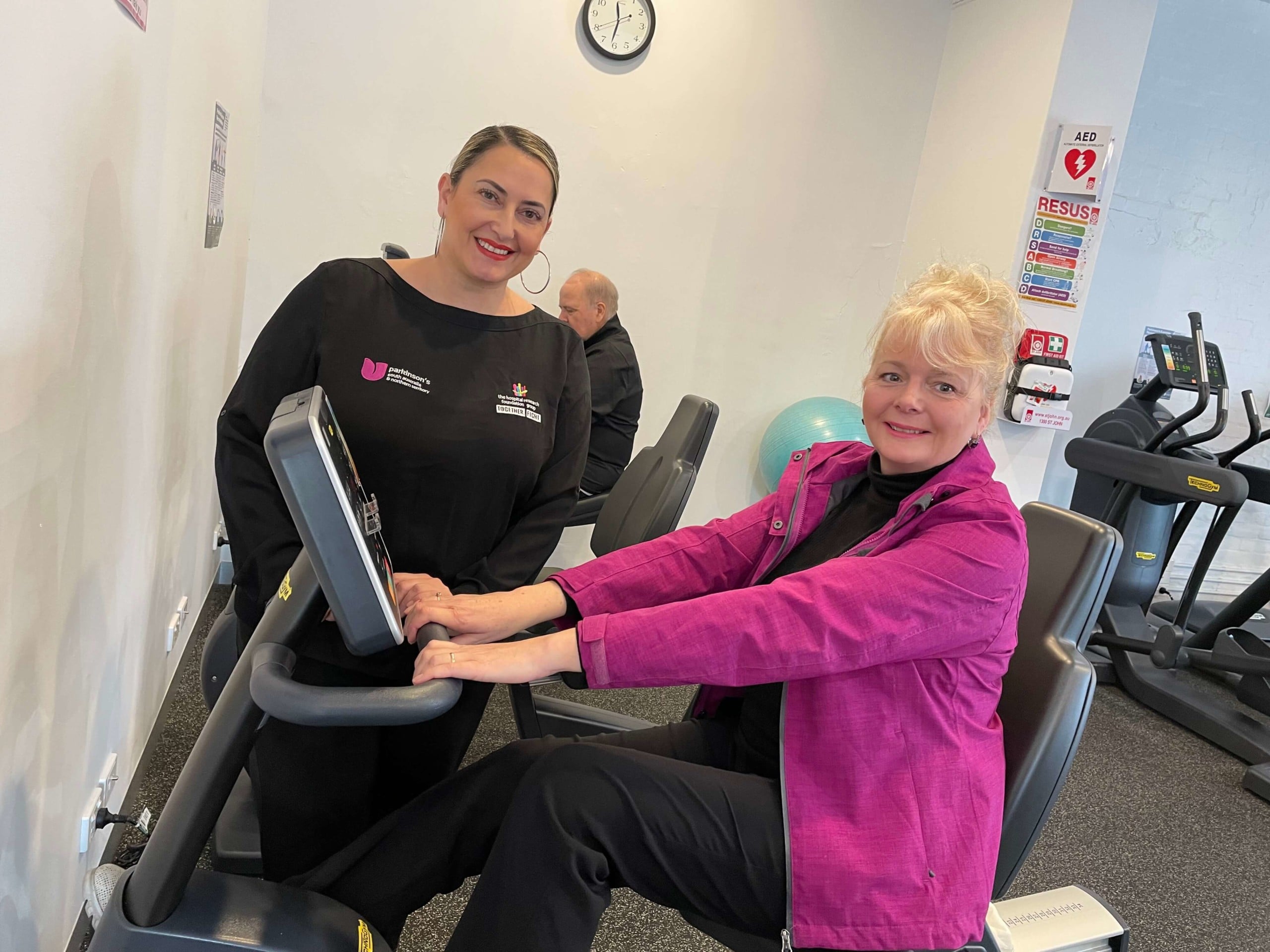 Two women smiling, one on an exercise bike