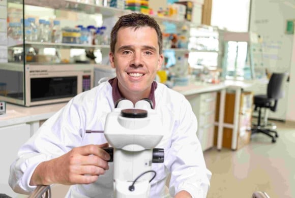 A male researcher wearing a white lab coat looking into a microscope