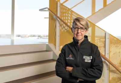 A blonde woman wearing a black jacket stands in a yellow stairwell