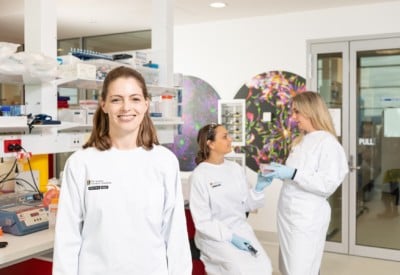 Three researchers in a lab, one is looking at the camera and two are in the background