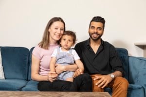 Adela with her husband Shah and son Amir smile at the camera, sitting on their couch