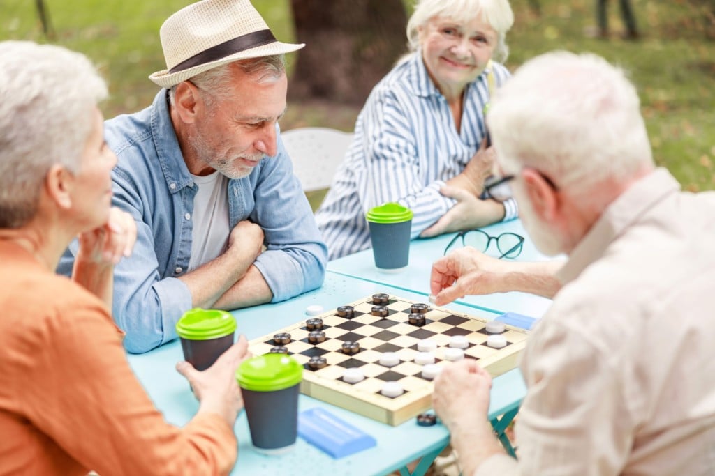 A group of four people sit playing chess, smiling at the camera. They are outside with a coffee in front of them.