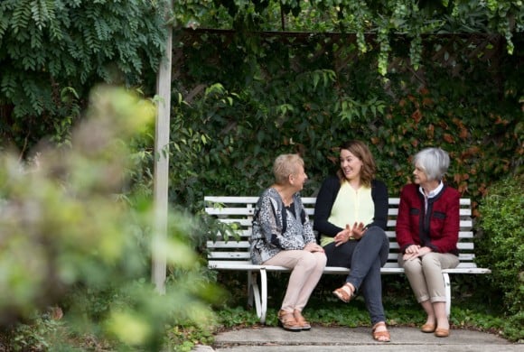 Three women sit on a bench surrounded by trees