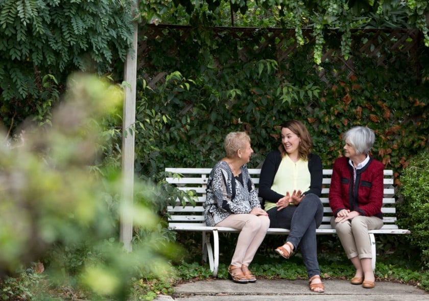 Three women sit on a bench surrounded by trees