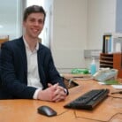 Dr Chris Belder sitting at a corner desk smiling to camera