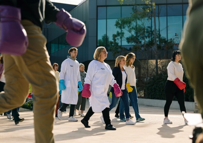 A group of people wearing white lab coats and coloured boxing gloves walk past the camera with fierce expressions