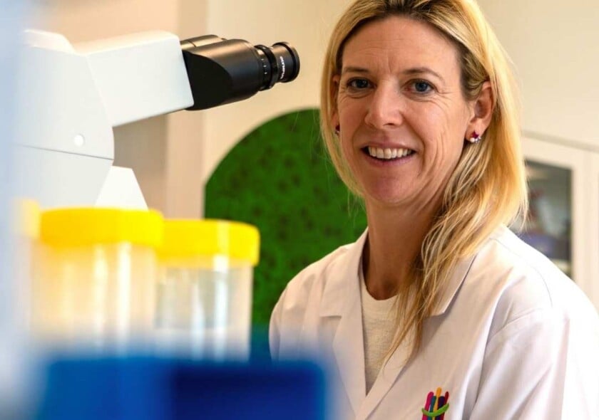 A woman wearing a lab coat smiles at the camera in front of a microscope