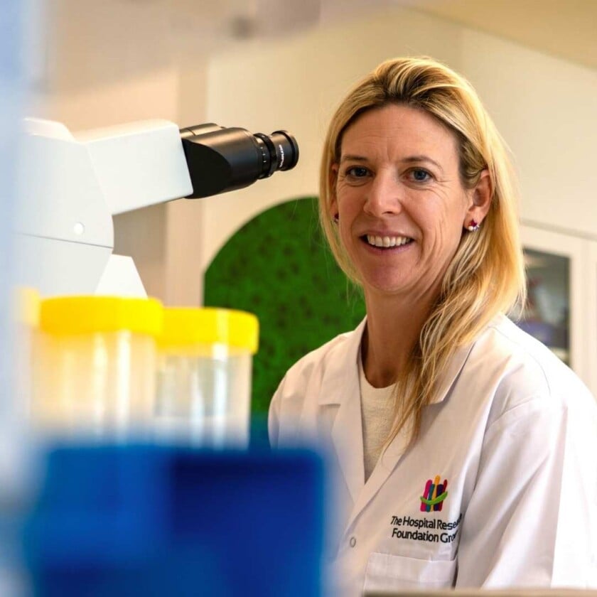 A woman wearing a lab coat smiles at the camera in front of a microscope