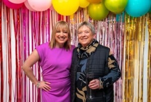 Jess and her mum smile at the camera in front of colourful streamers and balloons.