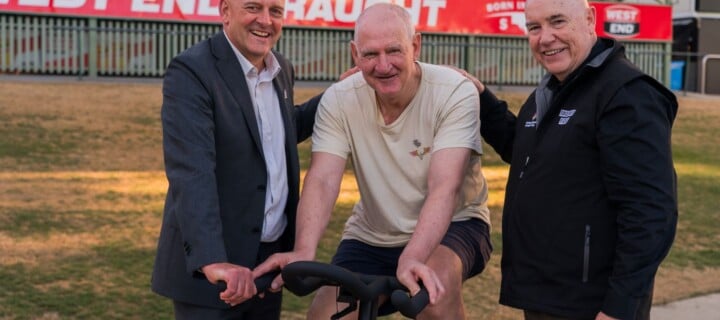 SANFL CEO Darren Chandler, Pedal to Fight Parkinson's ambassador and SA football legend Mark Mickan, and The Hospital Research Foundation Group CEO Professor Paul Flynn in front of the old scoreboard at Adelaide Oval.