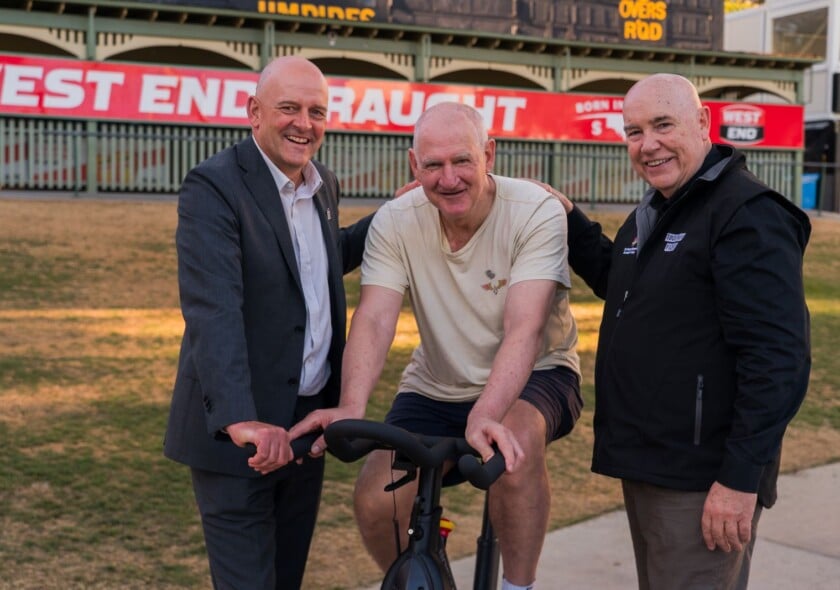 SANFL CEO Darren Chandler, Pedal to Fight Parkinson's ambassador and SA football legend Mark Mickan, and The Hospital Research Foundation Group CEO Professor Paul Flynn in front of the old scoreboard at Adelaide Oval.
