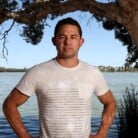 Phil Britten stands with hands on hips. Phil is wearing a white T-shirt. Behind Phil is a body of water.