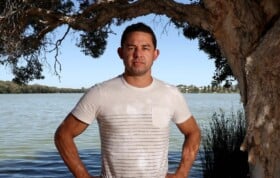 Phil Britten stands with hands on hips. Phil is wearing a white T-shirt. Behind Phil is a body of water.